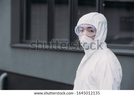 asian epidemiologist in hazmat suit and respirator mask looking at camera while standing on street Royalty-Free Stock Photo #1645012135