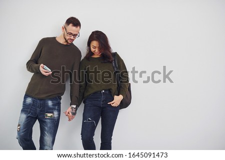 Cheerful multi ethnic couple with backpack and phone standing together indoors in the studio against white background.