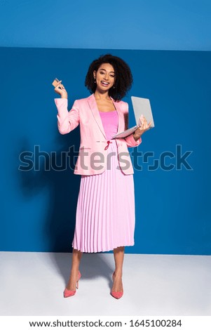 elegant african american businesswoman holding credit card and laptop on blue background