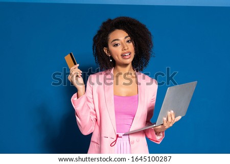 elegant african american businesswoman holding credit card and laptop on blue background
