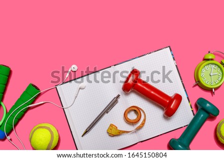 Healthy lifestyle concept with empty exercise book for text, fitness and sport. Tennis ball with dumbbells and green alarm clock isolated on pastel pink background.