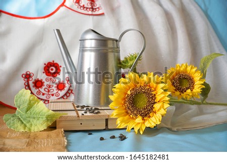 Still life, vintage style in ethno style, sunflowers, a metal watering can stand on a wooden stand against the background of an embroidered bright pattern.                             