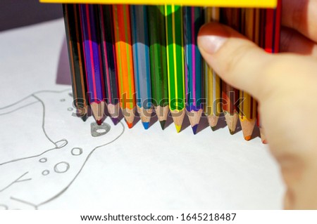 Children's hand holds color pencils on a white paper with a pattern. The child paints a picture. Children's creativity. Painting. Copy space
