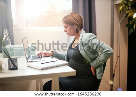 Cute caucasian pregnant businesswoman sitting on chair in office, holding back and using laptop.