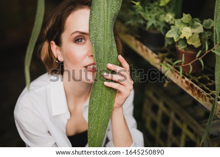 Young beautiful caucasian woman in glass greenhouse among colorful greenery leaves and flowers. Art portrait of a girl wearing a shirt. Gardener, nature lover, inspiration concept.