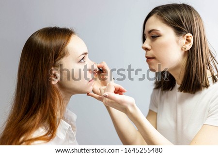 Skillful visagiste in white t-shirt is holding brush, applying make-up for young curly model while posing sideways against gray studio background. Fashion and beauty concept. Close up, copy space