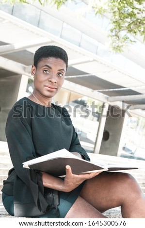 Pensive positive black woman studying album outside. African American lady sitting on outdoor stair, holding photo book and looking at camera. Book reader concept