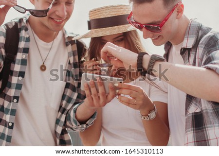 Youth Selfie And Chooses The Best Photo On The Phone Outdoor. Portrait Of Two Guys In Sunglasses And A Girl In A Straw Hat On A Summer Day. Warm Toned.