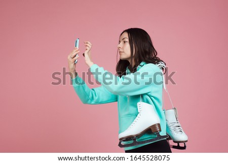 Figure skating, Hobbies and a healthy active lifestyle. A cheerful girl in a bright hoodie with skates on her shoulder is preparing to go to the skating rink. Uses a smartphone to take a selfie