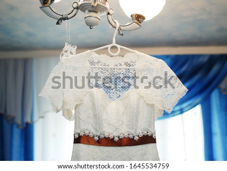 A white wedding dress with a brown satin ribbon at the waist hangs on a chandelier in the living room. Clothing for the bride