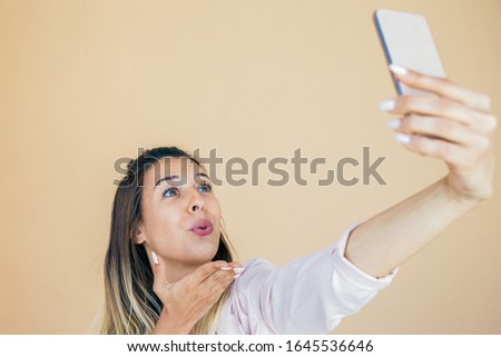Joyful excited lady holding cellphone in outstretched hand and sending air kiss to screen. Happy young woman taking selfie and posing. Self portrait concept