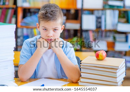 A boy sits at a table in a library with a stack of books thinking over a blank paper sheet.