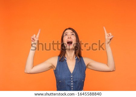 Wow look there, advertising area! Surprised happy brunette girl in dress looking astonished and pointing up copy space on orange background, showing empty place above head for promotion. studio shot