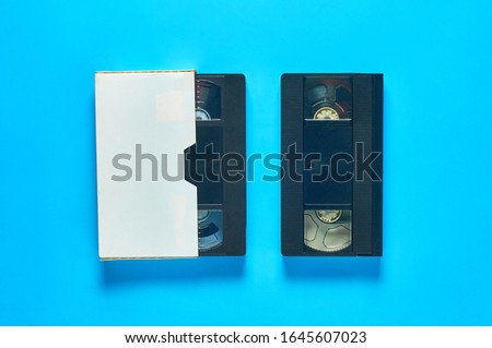 Black old vhs video cassette in white cardboard packaging on blue desk. Concept of 90s. Top view. Close-up