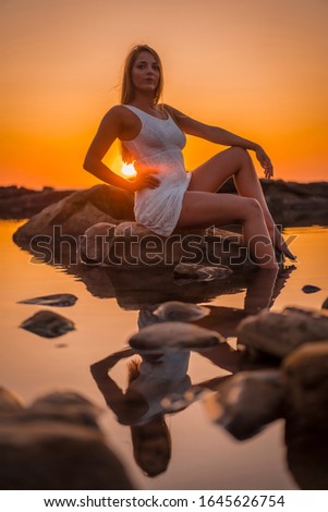 Lifestyle, a young blonde reflected in the water some rocks with a white dress on the coast of Basque Country, in an orange sunset