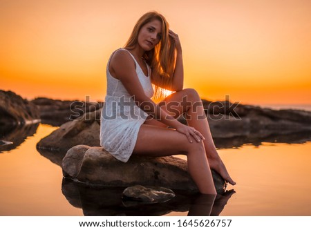 Lifestyle, a young blonde reflected in the water some rocks with a white dress on the coast