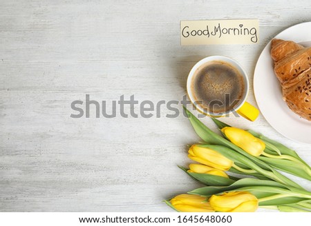 Delicious coffee, croissant, beautiful flowers and card with GOOD MORNING wish on white wooden table, flat lay. Space for text
