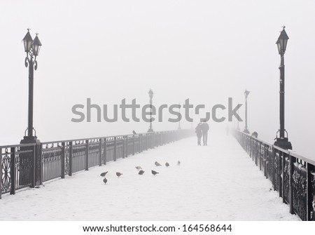 Bridge city landscape in foggy snowy winter day - walking couple, lanterns and doves flock - Ukraine, Donetsk