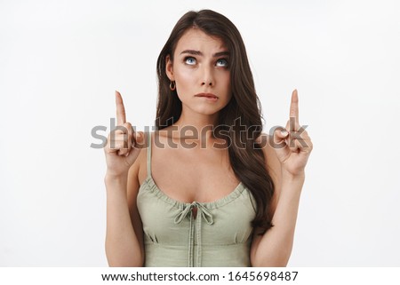 Temptation concept. Alluring indecisive brunette woman biting lip overwhelmed, looking and pointing up, tempting try something delicious, restrain herself but really want, white background