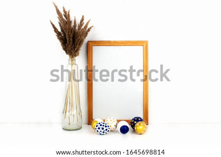 Empty frame mockup with copy space, modern golden, blue and white easter eggs, trendy dry flowers in glass bottle on white background. Happy easter celebration concept. Banner template. Stock photo.