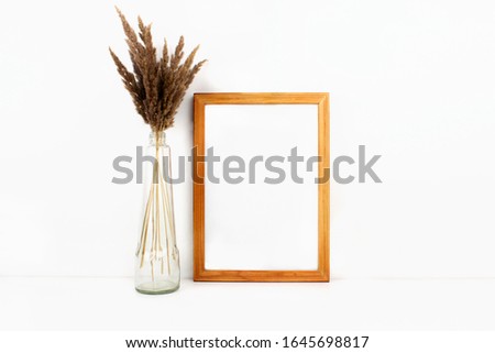 Wooden empty frame mockup with copy space, trendy dry flowers in glass bottle on white background. Interior vertical poster mock up in minimal style. Web, social media banner template. Stock photo.