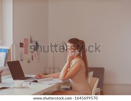 Attractive woman sitting at desk in office, working with laptop computer, holding document