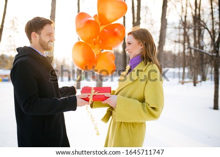 saint valentines day, love concept. young caucasian couple celebrating. handsome bearded guy give a gift to lady in coat outdoors, couple stand with red air balloons outdoors
