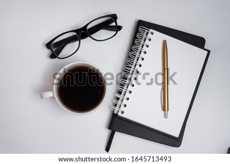 The begining of the work day. Morning at the office. Student workplace. Cup of coffee, black notebook, pen, white sheet with empty place for text and glasses on a white background. View from above.