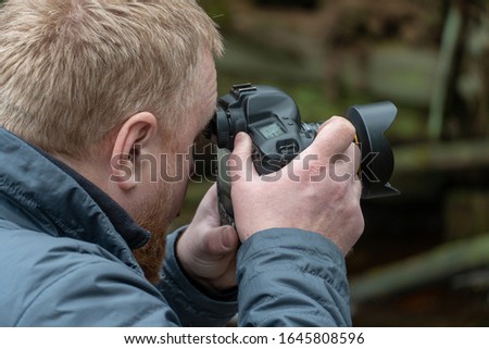 Photographer in the forest. Professional camera in the hands. Photographing nature and landscape. Fashionable young guy with a beard holds a camera.