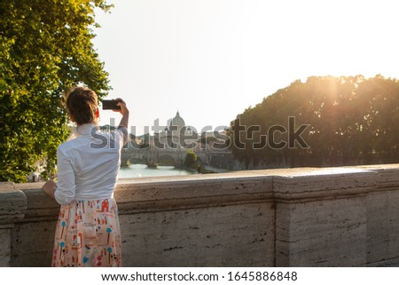 Young woman looking at St Peter's basilica from a bridge on tiber river at sunet, taking pictures with smartphone. Panoramic view cityscape of Rome, Italy.