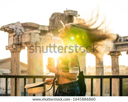 Beautiful young woman shaking long blond hair at Roman Forum at sunrise. Happy smiling tourist with vintage camera. Lens flair. Rome, Italy