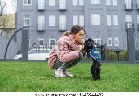 Attractive lady playing with her dog sitting on the lawn against the background of architecture, wearing a pink coat. Cute woman having fun with a dog on a walk. Weekend with your pet.