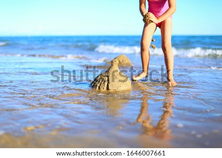 Legs of cute little girl spend holidays in pink one-piece swimsuit,builds sand castle,plays in the water on the shore of blue sea. Vacation with kids on beach on warm sunny summer day before sunset.
