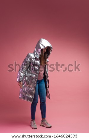 Trendy autumn and winter clothing, Studio shot on a pink background, copy space. A cool girl in a shiny silver down jacket and a knitted hat