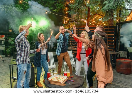Caucasian friends having garden party outside, they dancing to the Dj's music with bengal lights in house backyard at summer night.