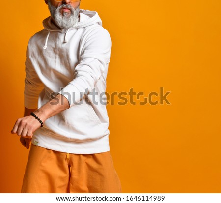 gray-haired, bearded, mature male in a white sweatshirt, pants and sunglasses, bracelets. There is dancing posing on an orange background. joyful old lifestyle