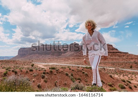 A curly haired blonde man standing around the famous Buttes of Monument Valley from Arizona, USA, wearing white cotton shirt and white pants, black shoes