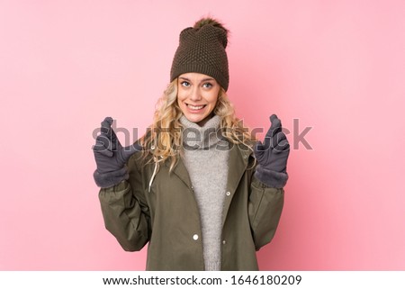 Young girl with winter hat isolated on pink background with fingers crossing