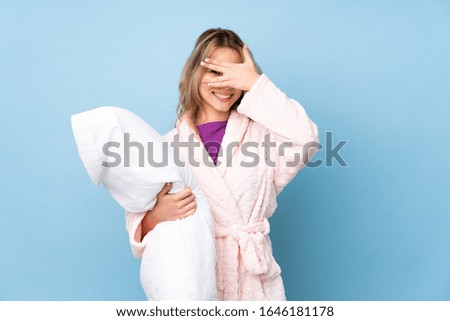 Teenager Russian girl in pajamas isolated on blue background covering eyes by hands and smiling