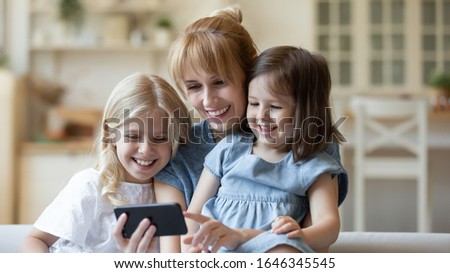 Happy young Caucasian mom and little preschooler daughters relax at home watching video on smartphone together, smiling mother or nanny rest with small little girls children laugh have fun using cell