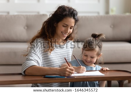 Front view joyful young mommy holding colored pencils, drawing cute pictures with adorable small child daughter, sitting at coffee table in living room. Happy babysitter educating developing kid. Royalty-Free Stock Photo #1646345572