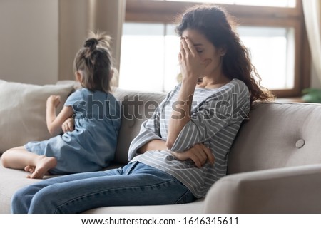 Unhappy young mother touching forehead, feeling tired of bad daughter's behavior at home. Offended little child girl sitting on different side on couch, ignoring sad frustrated mother in living room. Royalty-Free Stock Photo #1646345611