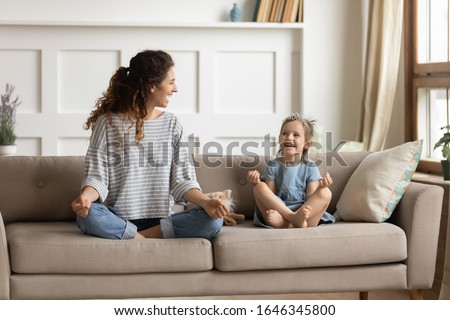 Full length front view smiling young curly mother sitting on comfortable couch with cute playful little preschool daughter in lotus position. Happy mommy practicing yoga exercised with small child. Royalty-Free Stock Photo #1646345800