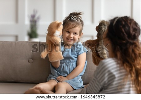 Happy small preschool kid girl sitting on couch, wearing crown, playing with nanny. Playful little child daughter holding fluffy toy, having fun with devoted young mommy at home, childcare concept. Royalty-Free Stock Photo #1646345872