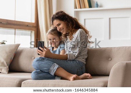 Happy beautiful young mother resting on couch with little preschool daughter, showing funny cartoons on mobile phone. Smiling affectionate family of two posing for smartphone selfie shot on sofa.