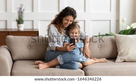 Front view happy pretty mommy cuddling small kid daughter, taking selfie shot on smartphone or playing online game at home. Loving young mother embracing little child, using mobile apps together.
