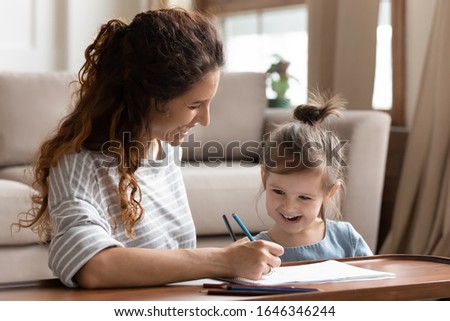 Head shot pretty happy woman engaged in early child education, teaching drawing little preschool girl at home. Smiling young mother helping small laughing adorable daughter painting picture in album.
