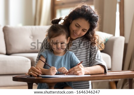 Pleasant young curly woman sitting at coffee table with preschool small kid girl, teaching helping drawing. Smiling little daughter holding pencils, coloring picture in paper album with pretty mother.
