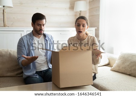 Frustrated husband and wife sit on couch in living room open cardboard package disappointed with internet online shopping, confused upset young couple unpack parcel confused with wrong order Royalty-Free Stock Photo #1646346541