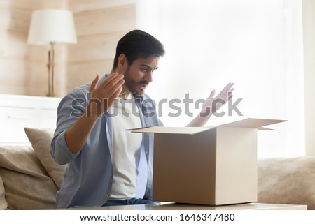 Frustrated Caucasian millennial male client open cardboard delivery parcel disappointed with wrong order, confused man customer unpack post shipping package upset with bad delivery service Royalty-Free Stock Photo #1646347480